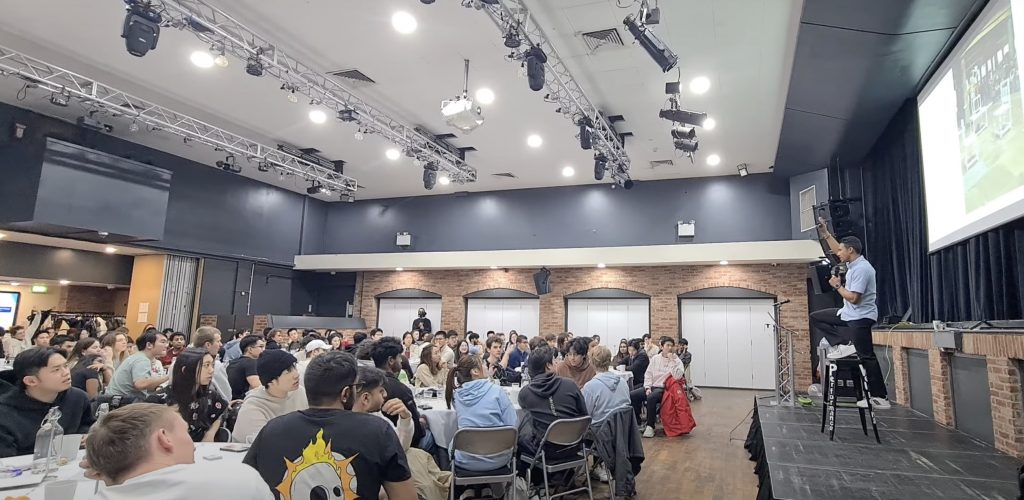 A well-being motivational speaker engages a large audience in a corporate event, addressing a diverse group seated at tables in a spacious venue.